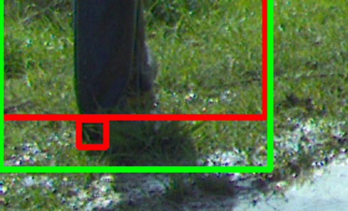 Zoomed view of frame 90 around the worker's feet. Green box is YOLO's predicted person at conf 0.846. Red boxes are the two ground-truth person components: the large silhouette (correctly covered by YOLO) and a small 22 by 22 pixel blob in the wet grass just below the boot, which is a labeling misfire rather than a real person or body part.